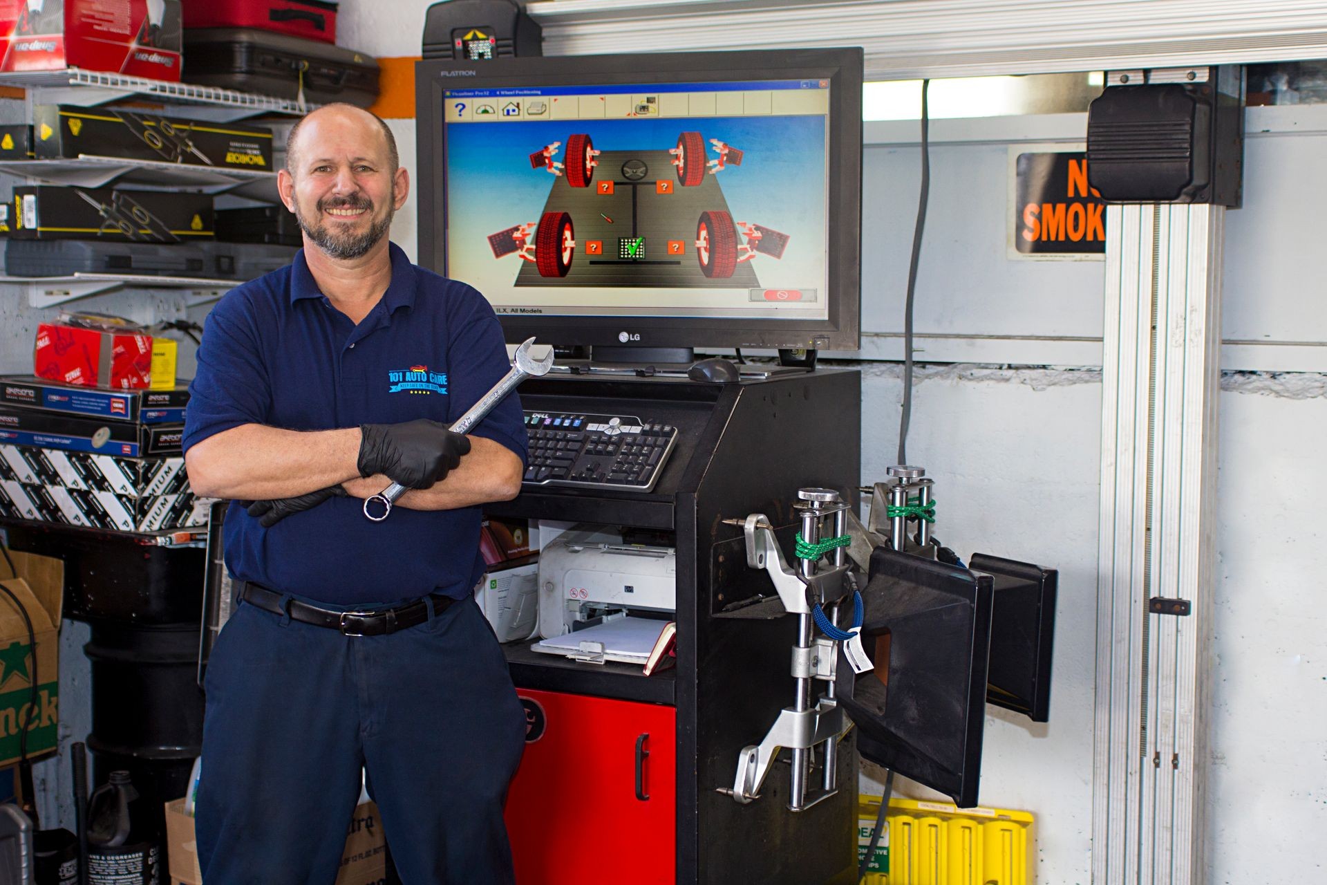 Smiling mechanic holding a wrench in an auto repair workshop with a computer displaying wheel alignment settings.