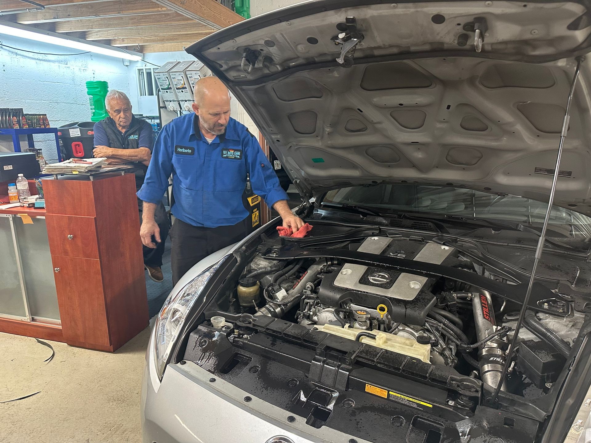 Mechanic in blue uniform working on a car engine while another man observes from behind a counter.